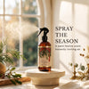 An amber spray bottle labeled Nutmeg & Cedar sits on a round pedestal in a sunlit room, with mist spraying from the nozzle. Text on the right reads 'SPRAY THE SEASON' and 'A warm festive scent.
