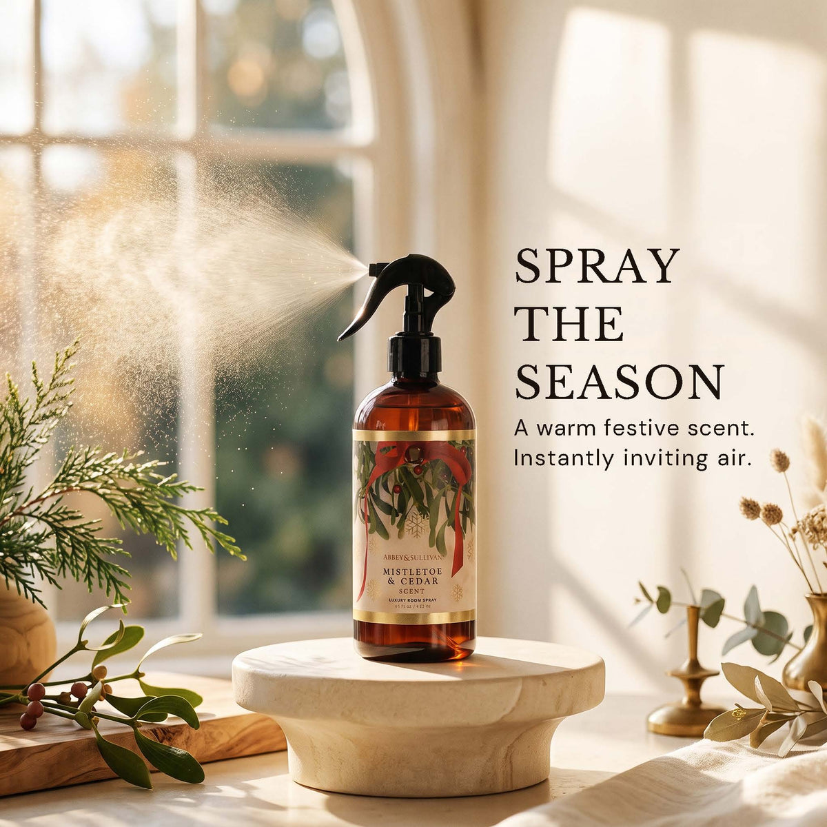 An amber spray bottle labeled Nutmeg & Cedar sits on a round pedestal in a sunlit room, with mist spraying from the nozzle. Text on the right reads 'SPRAY THE SEASON' and 'A warm festive scent.